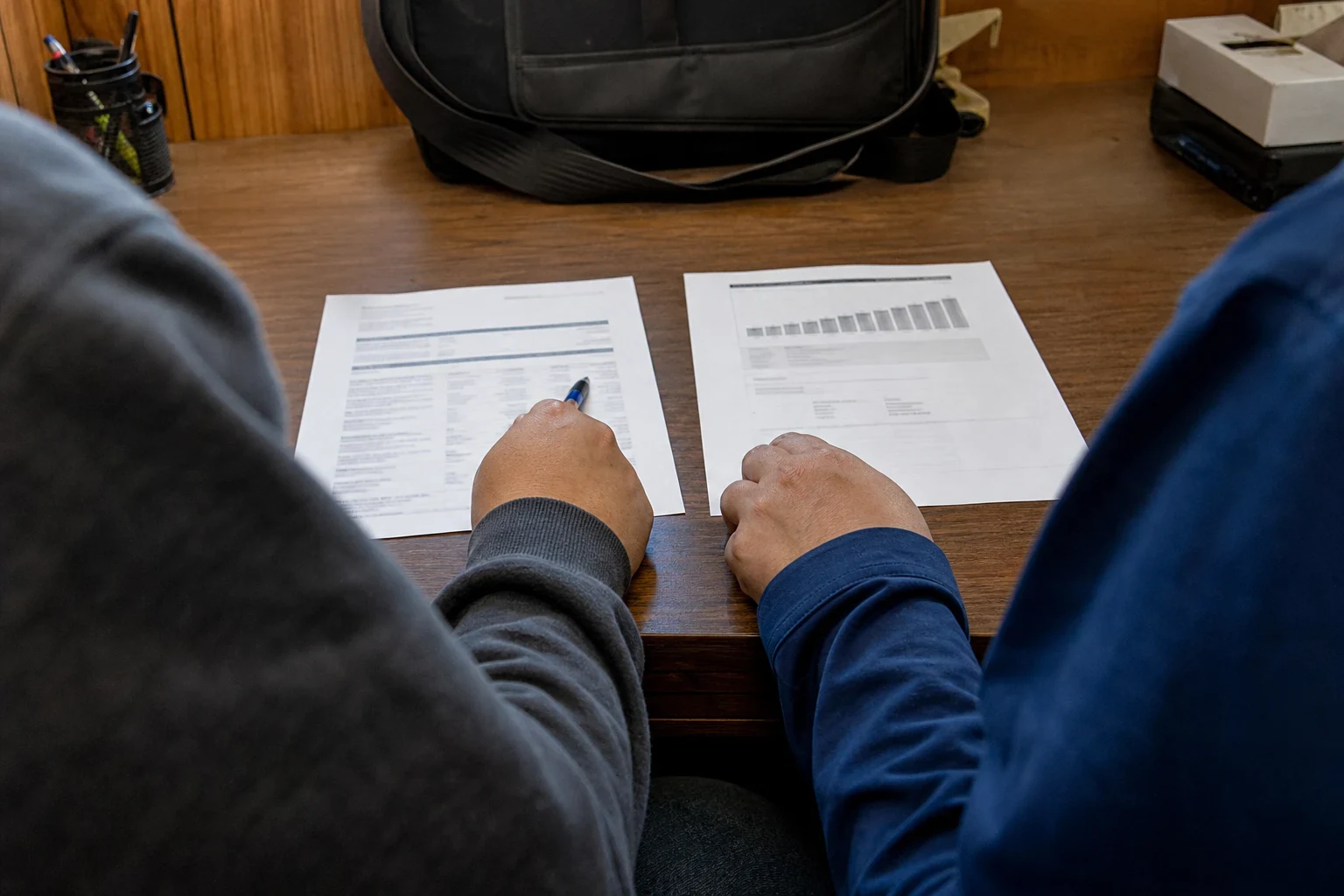 Over-the-shoulder view of two people at a wooden office desk, one pointing a pen at a statement beside a printed coverage chart, with a black bag and pen cup behind